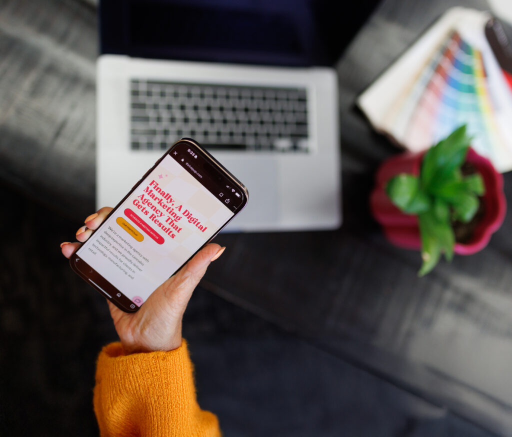 someone holding a phone showing a website with a laptop in the background